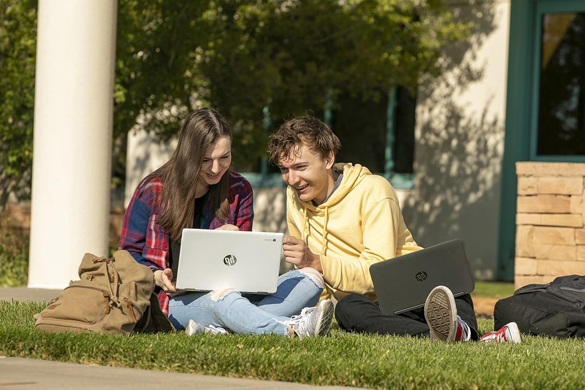 Two students sitting on the grass looking at a laptop laughing